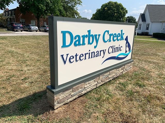 Sign for Darby Creek Veterinary Clinic; blue text on off-white background, stone base, grassy setting.