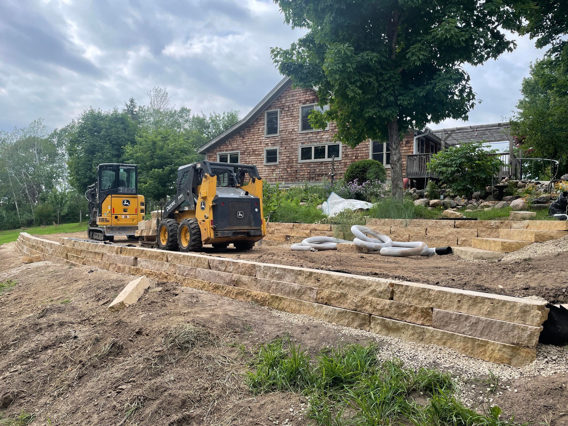 Construction site with retaining wall, two small tractors, and a house.