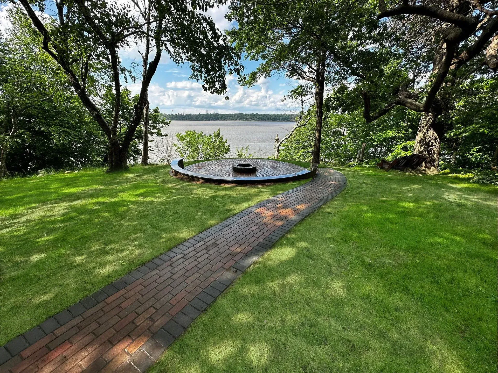 Brick path leads to a circular fire pit on a grassy bluff overlooking a body of water.