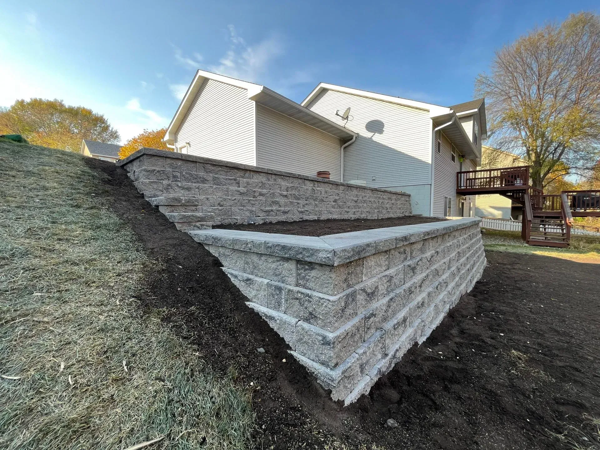 Two-tiered retaining walls made of gray blocks on a sloped yard next to a two-story beige house.