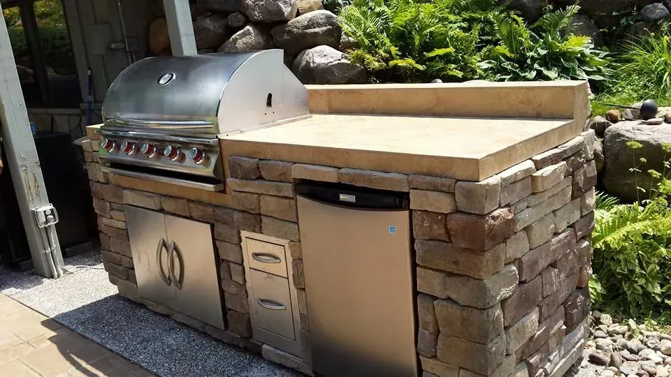 Outdoor kitchen with a stainless steel grill, fridge, and stone facade.