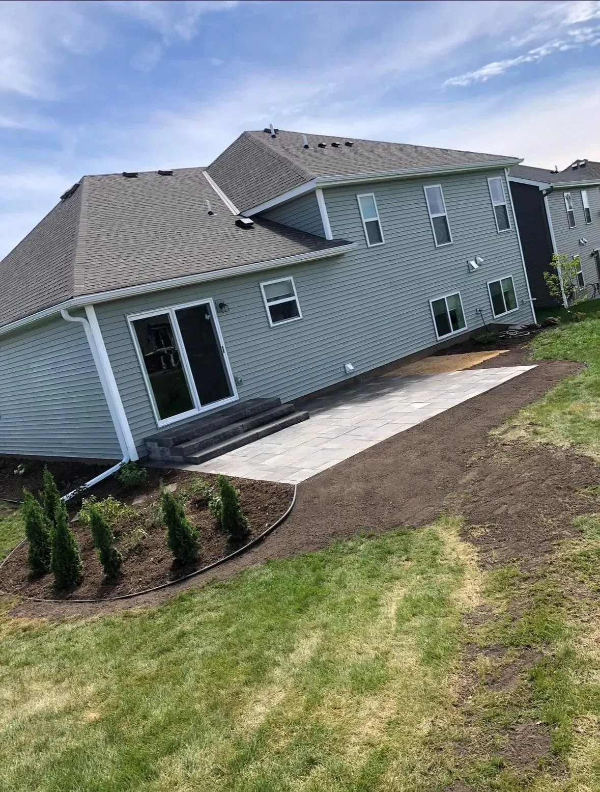 Backyard with light blue house, gray roof, concrete patio, and landscaping under a sunny sky.
