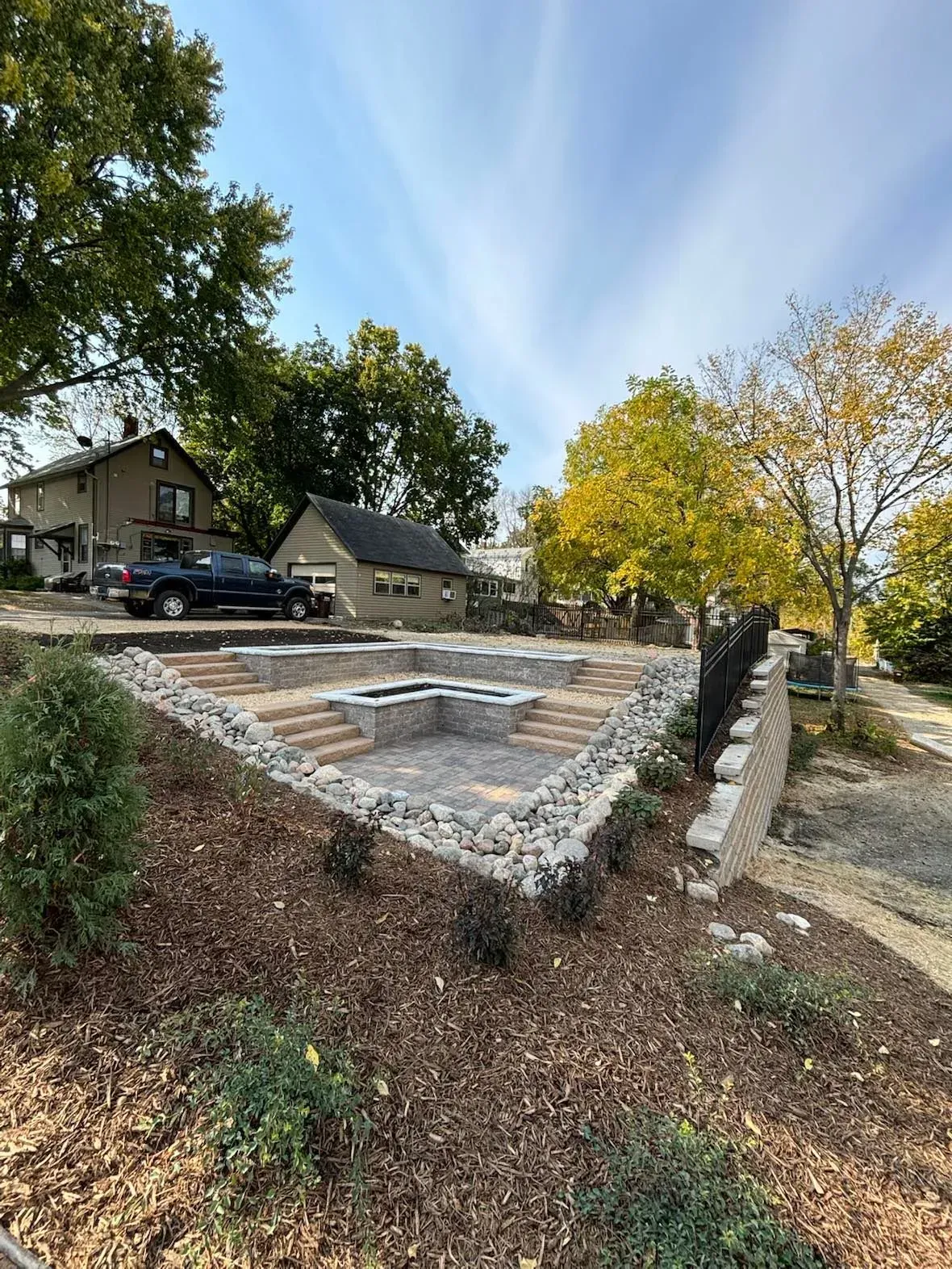 Landscaped area with retaining walls, stepping stones, and gravel. Buildings and trees in background.