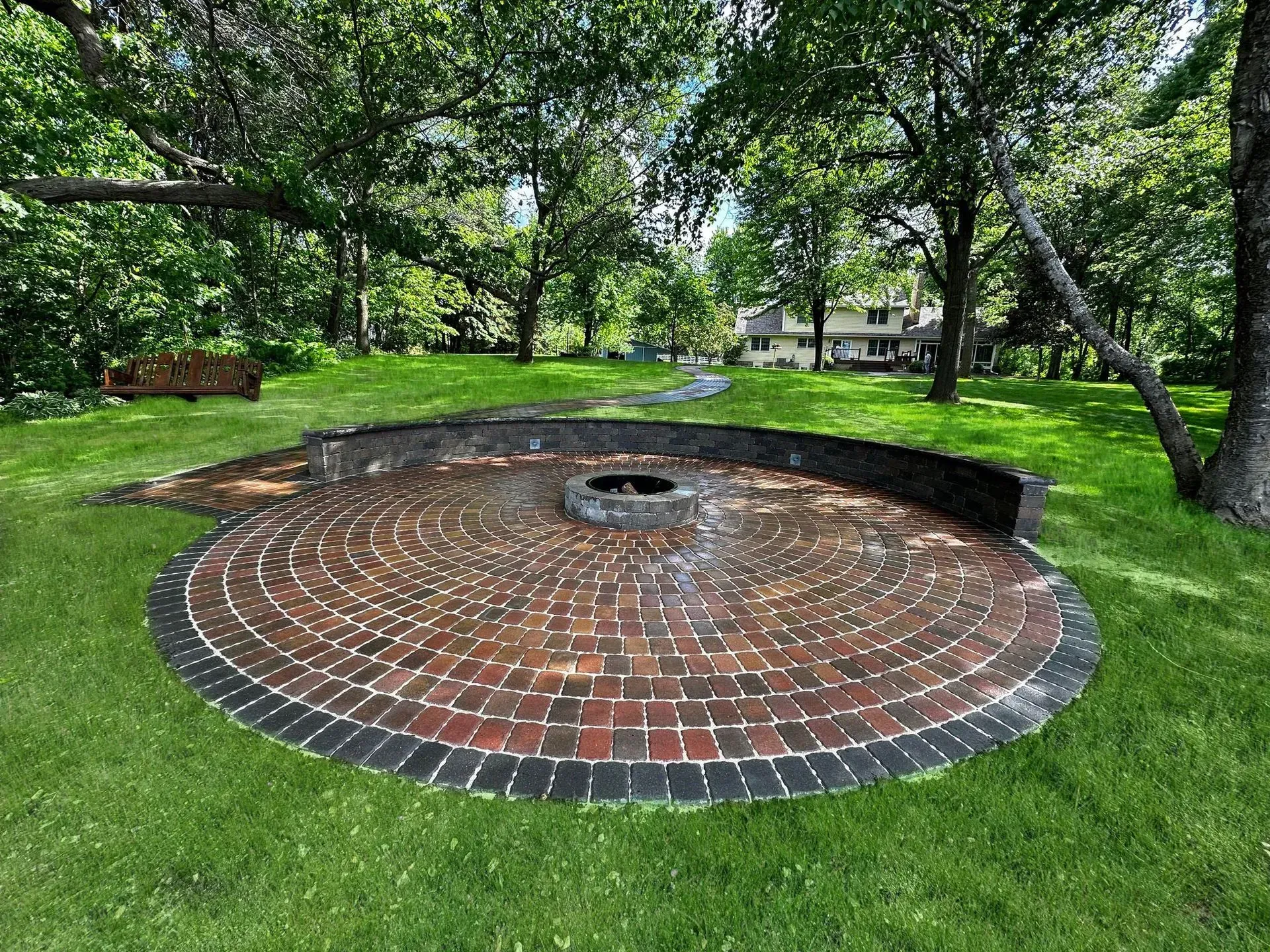 Circular brick patio with fire pit, surrounded by green grass and trees.