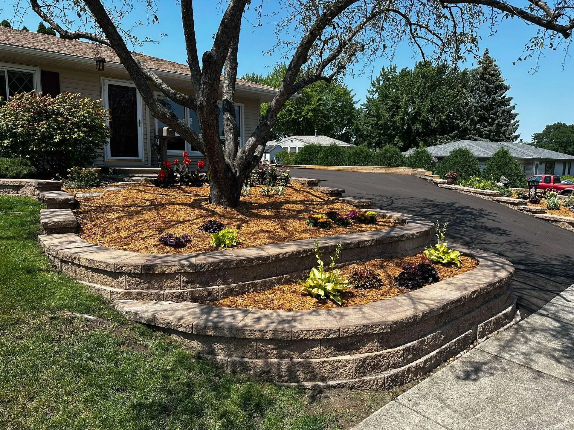 Multi-tiered retaining wall with mulch and plants surrounds a tree in front of a house.