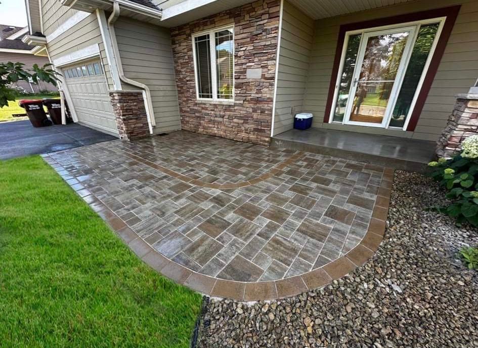 Curved brick patio beside a house with stone siding, bordered by grass and gravel.