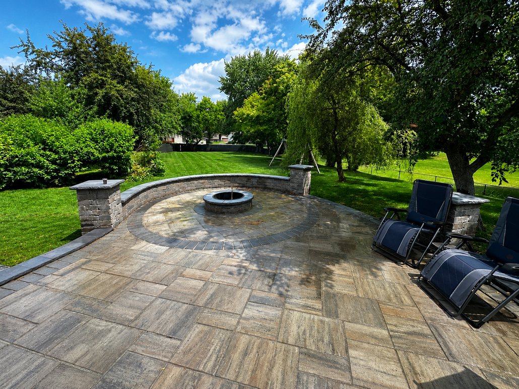 Stone patio with a central fire pit, lounge chairs, and a lush green garden under a blue sky.
