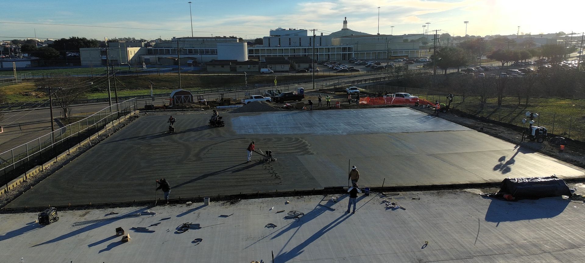 Construction workers pouring concrete on a large, rectangular surface. Buildings and sky in the background.