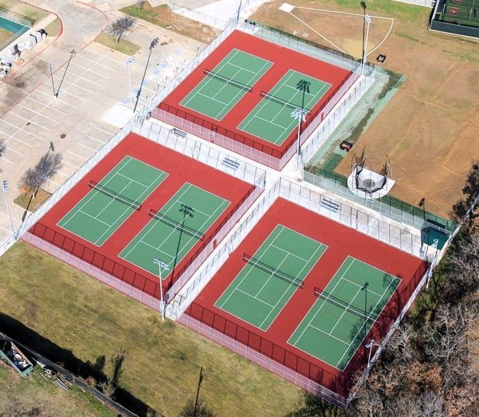 An aerial view of six outdoor tennis courts with green playing surfaces and red borders, arranged in three pairs.