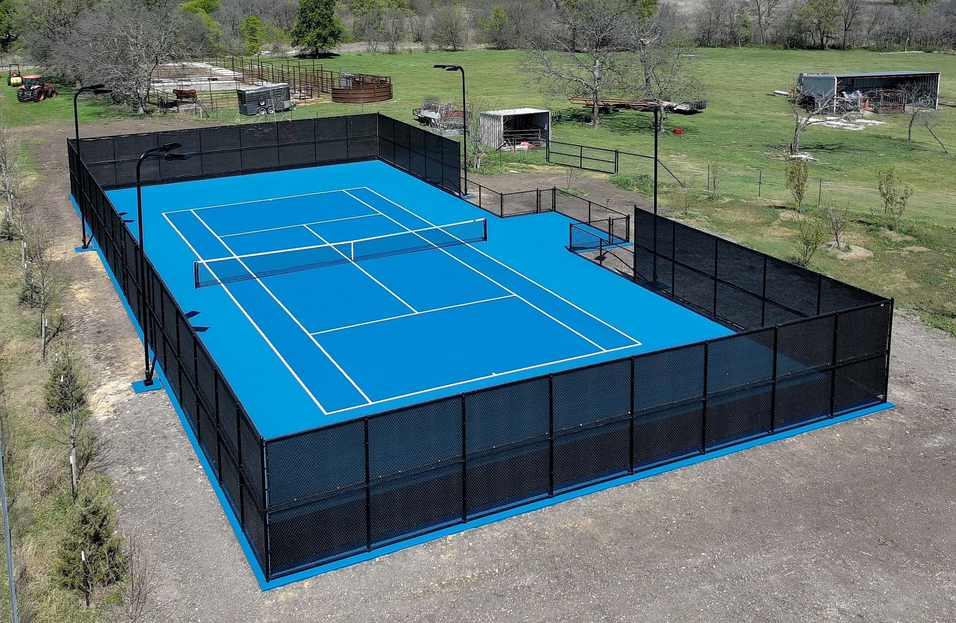A vibrant blue tennis court with black fencing in a rural setting.
