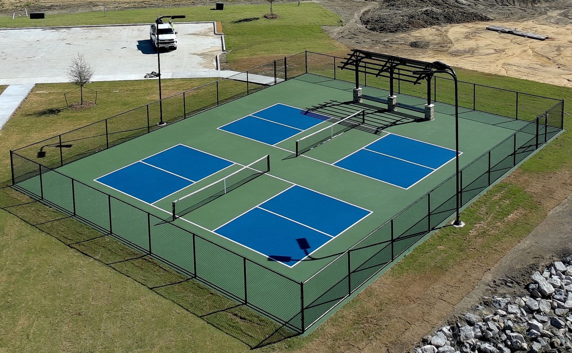 An aerial view of four blue and green pickleball courts enclosed by a black fence in a grassy outdoor area.