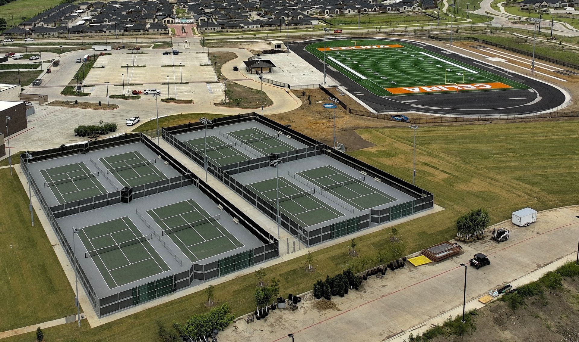 Aerial view of multiple blue tennis courts with black fencing. Several houses are visible in the background.