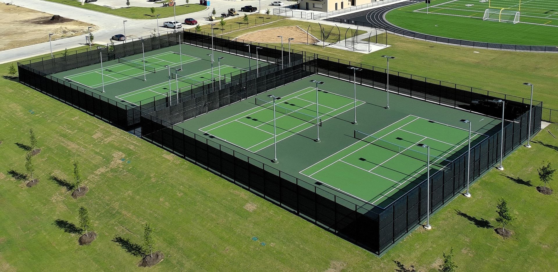 Aerial view of fenced outdoor tennis courts with green and blue surfaces beside a grassy field