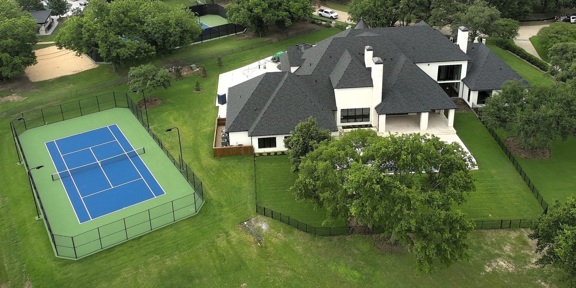 Aerial view of a house with a blue tennis court and surrounding green lawn and trees.
