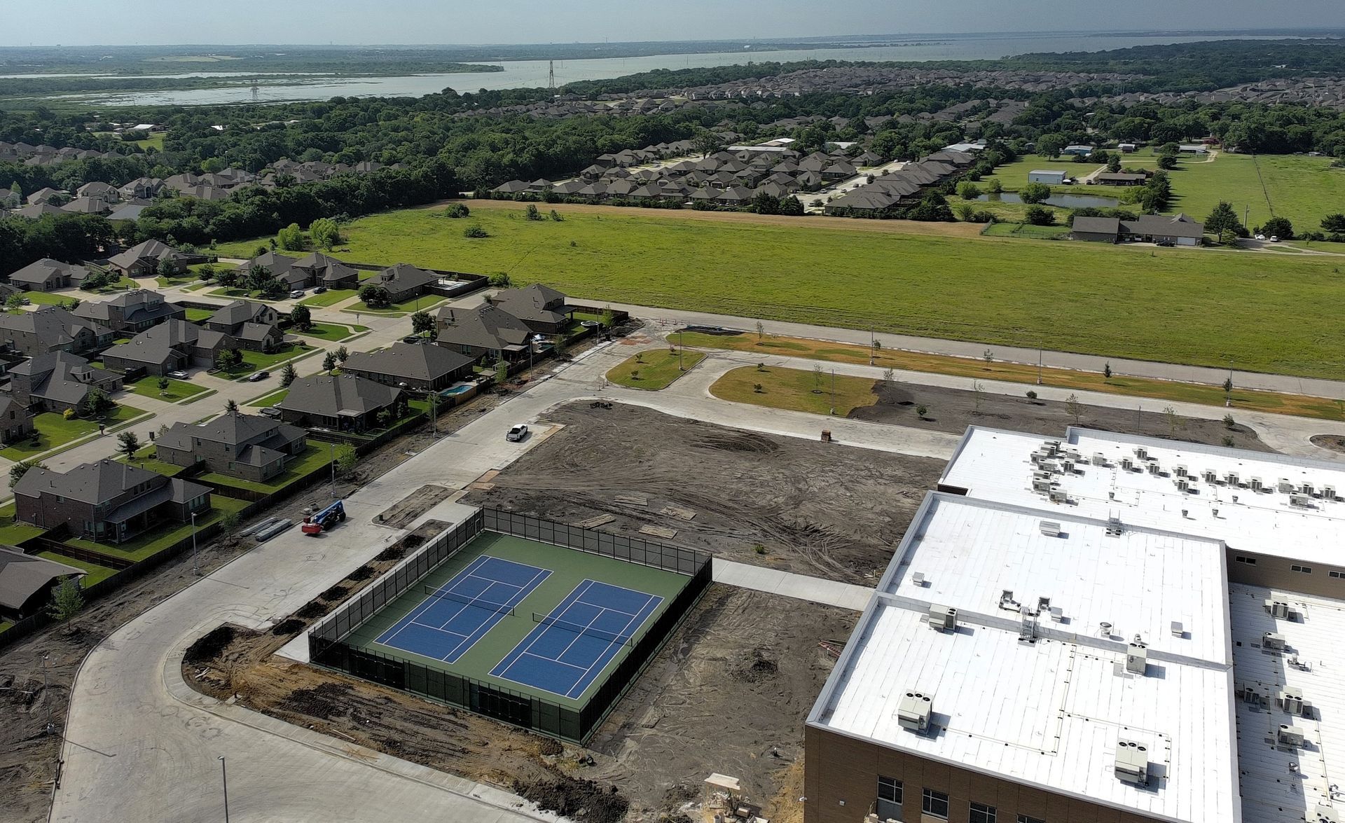 Aerial view of a suburban neighborhood with new construction, blue tennis courts, and a lake in the distance