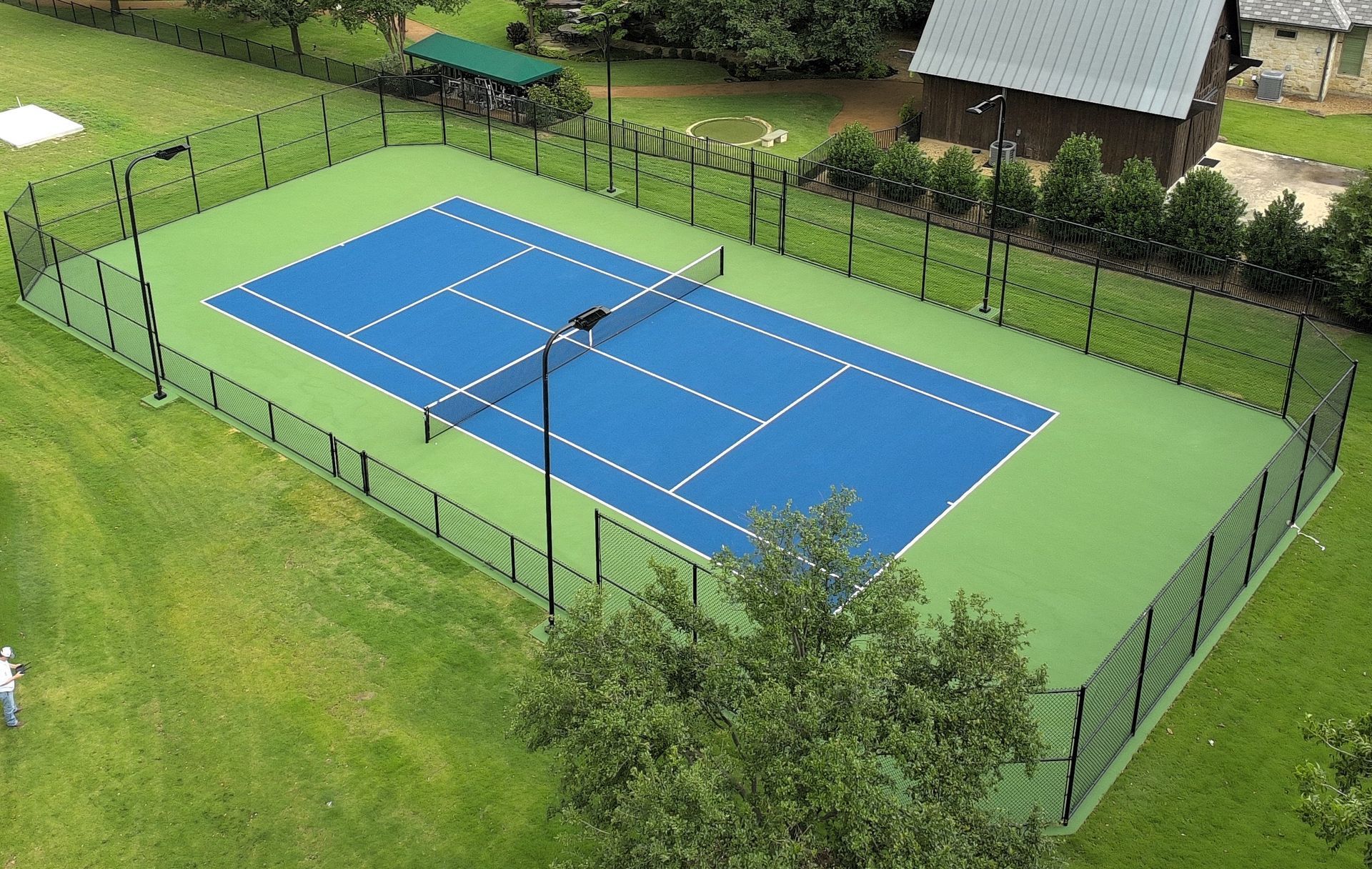 A fenced blue tennis court surrounded by green grass and trees, viewed from above.
