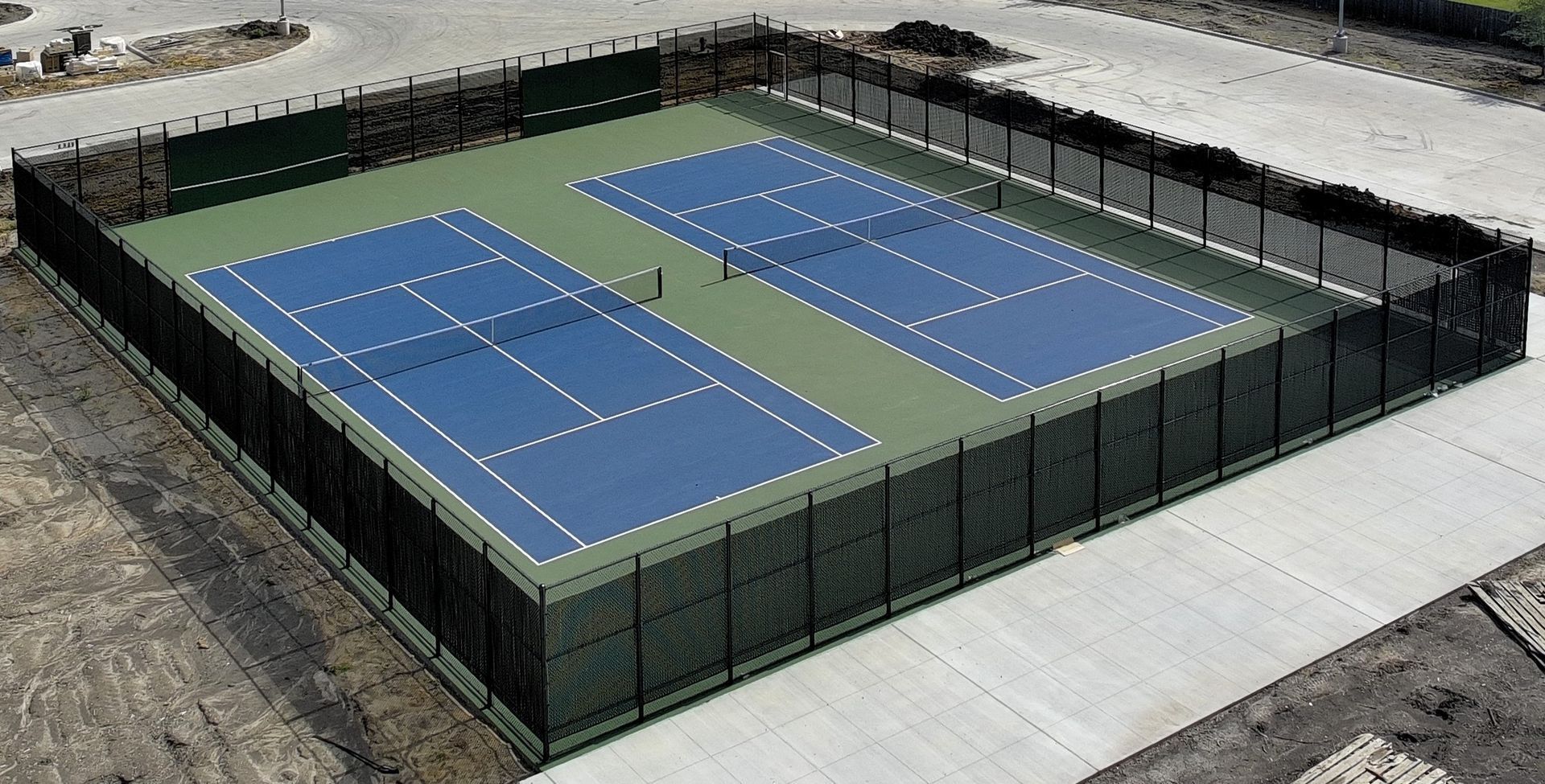 Aerial view of a fenced outdoor pickleball court with two blue playing surfaces on a concrete lot
