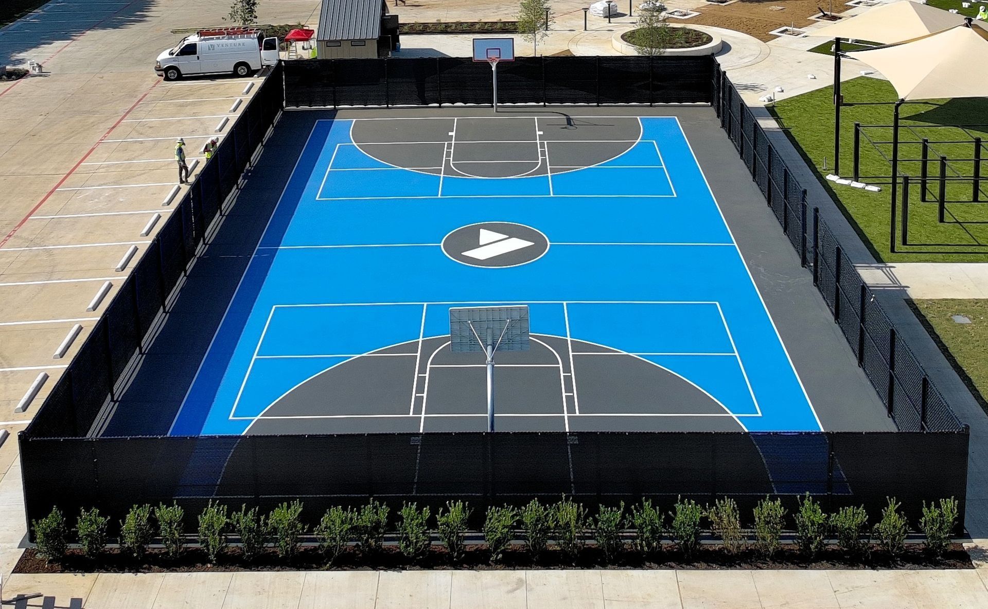 Aerial view of a fenced outdoor basketball court with bright blue and black pavement and white markings.