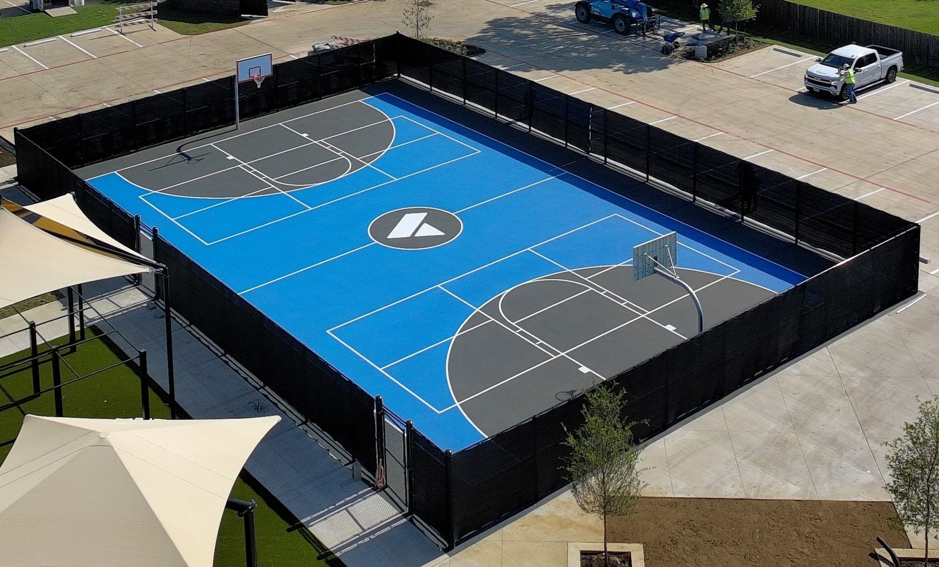 Aerial view of a blue outdoor basketball court with black fencing and surrounding pavement