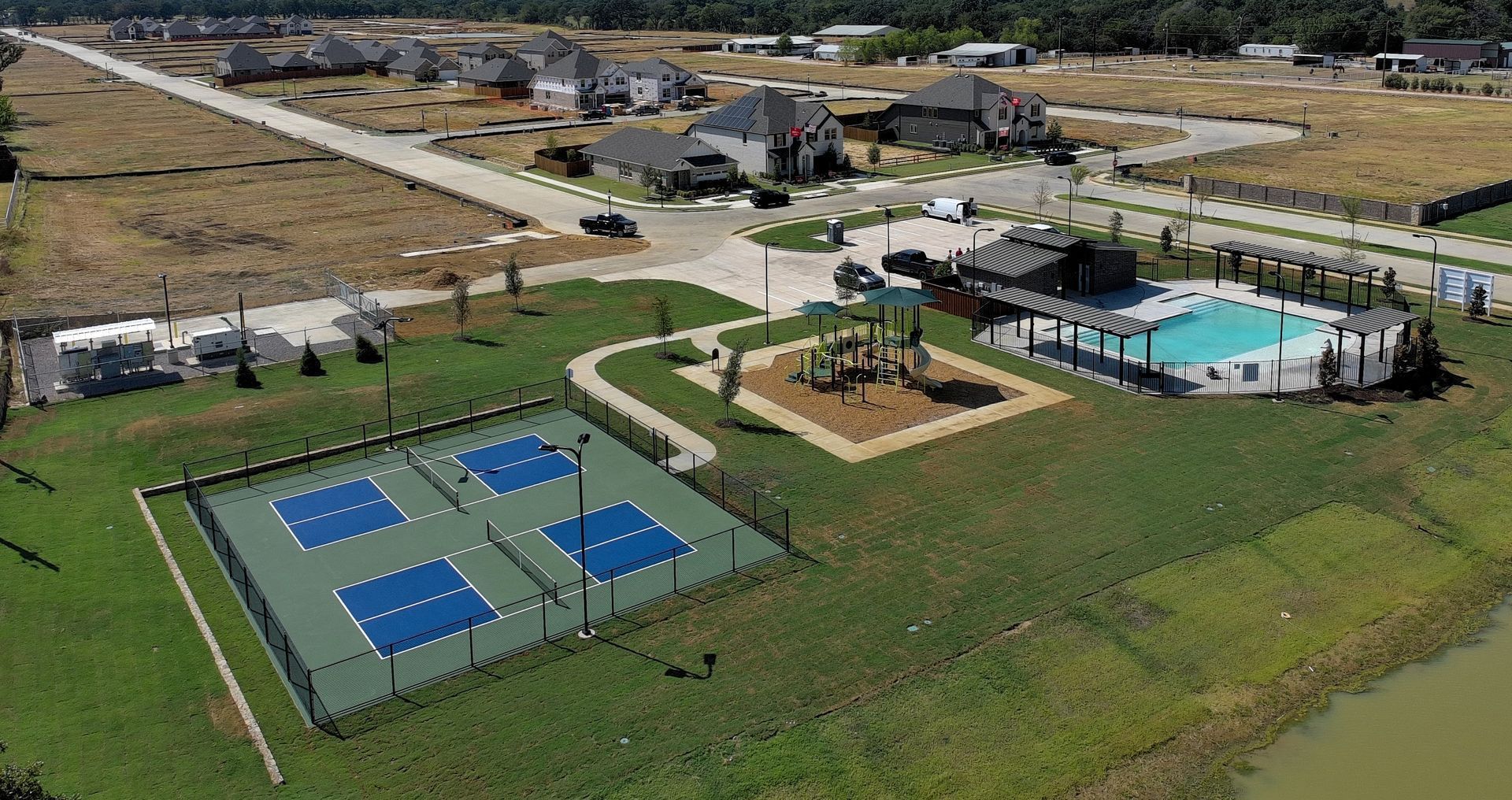 Aerial view of park with pickleball courts, a pool, and landscaped paths beside a grassy field