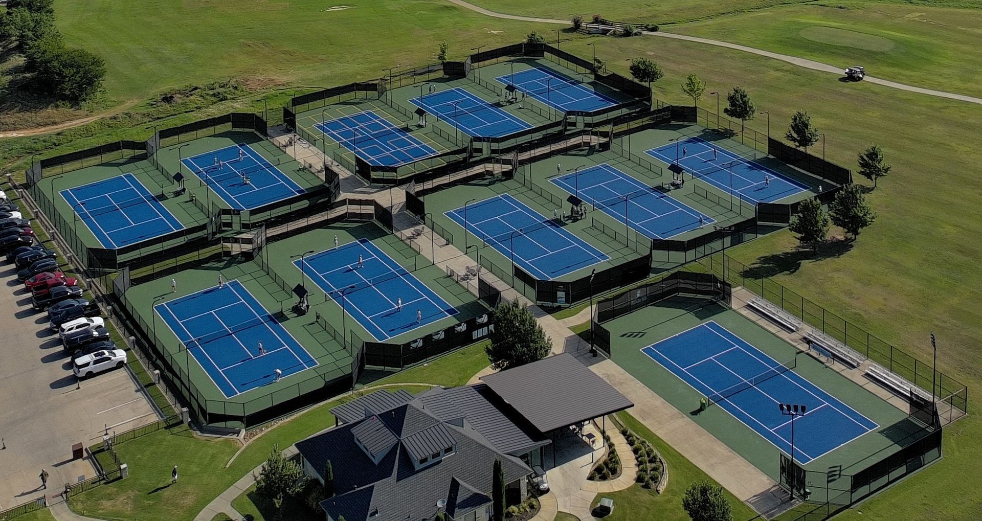 Aerial view of a tennis complex with many blue courts, surrounding grass, and a central clubhouse.