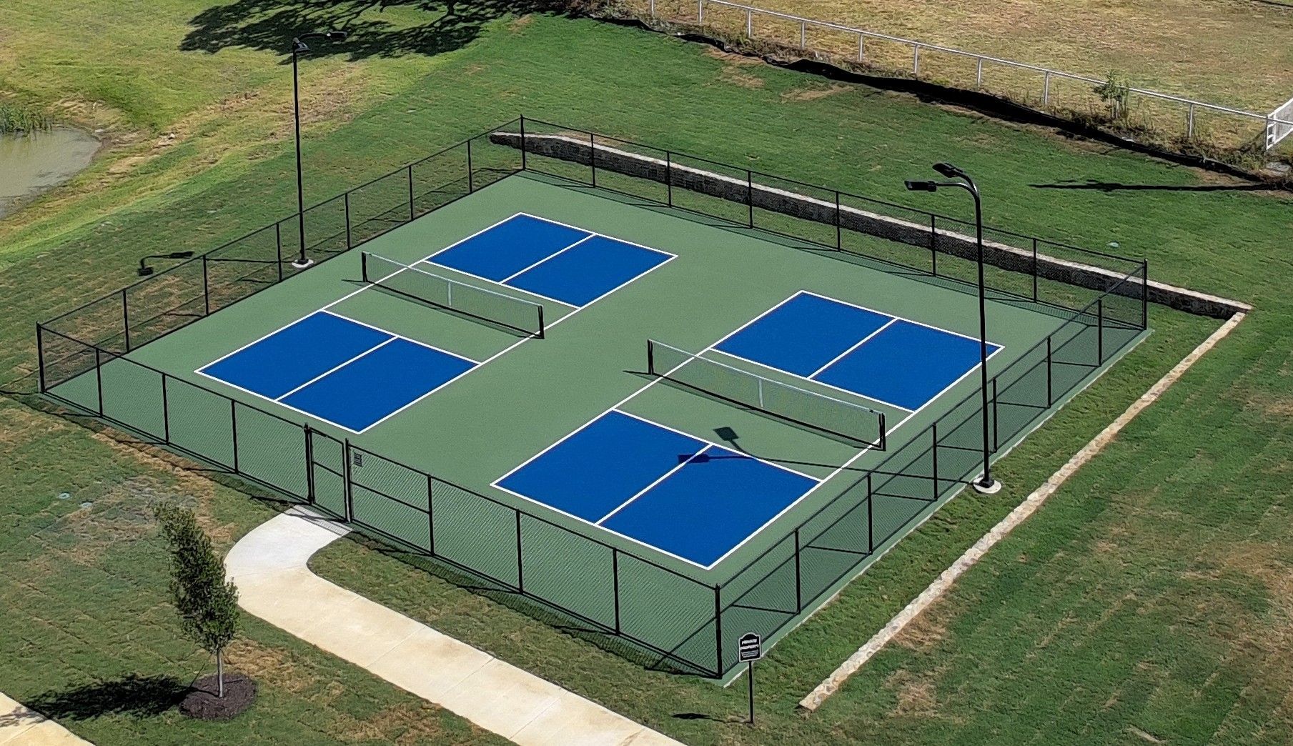 A fenced outdoor tennis/pickleball court with four blue playing areas on a grassy field.