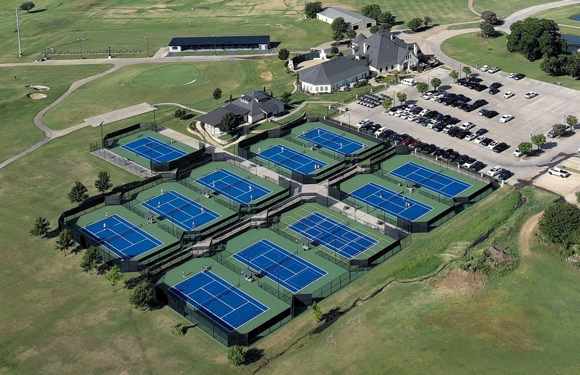 Aerial view of outdoor blue tennis courts beside a parking lot on a grassy sports complex