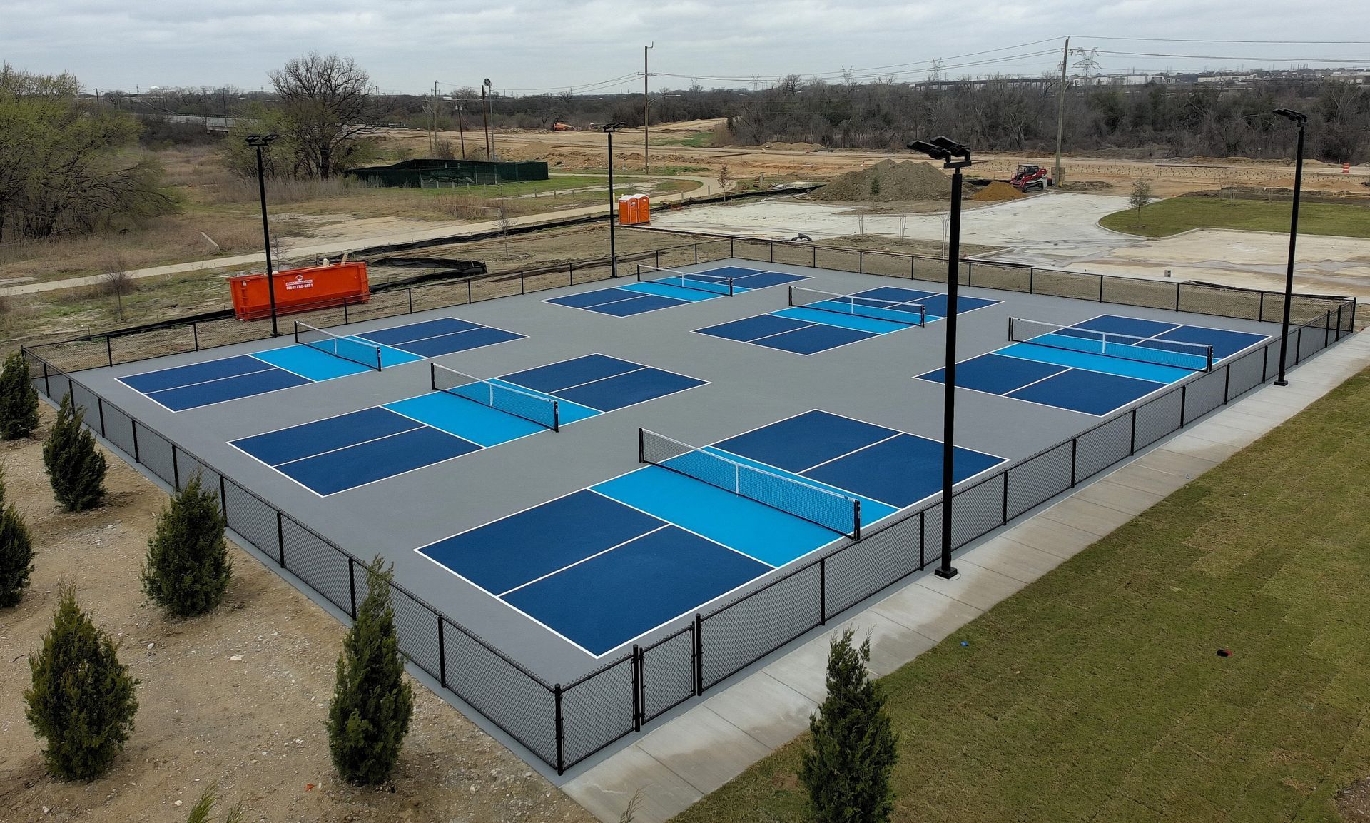 Outdoor pickleball courts, blue and gray surfaces, fenced in.