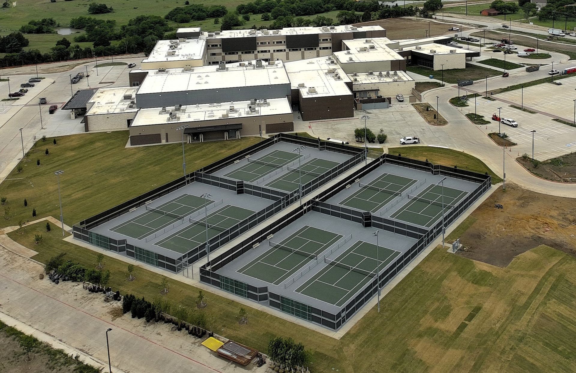 Aerial view of a tennis facility with multiple green and gray courts beside a large building.