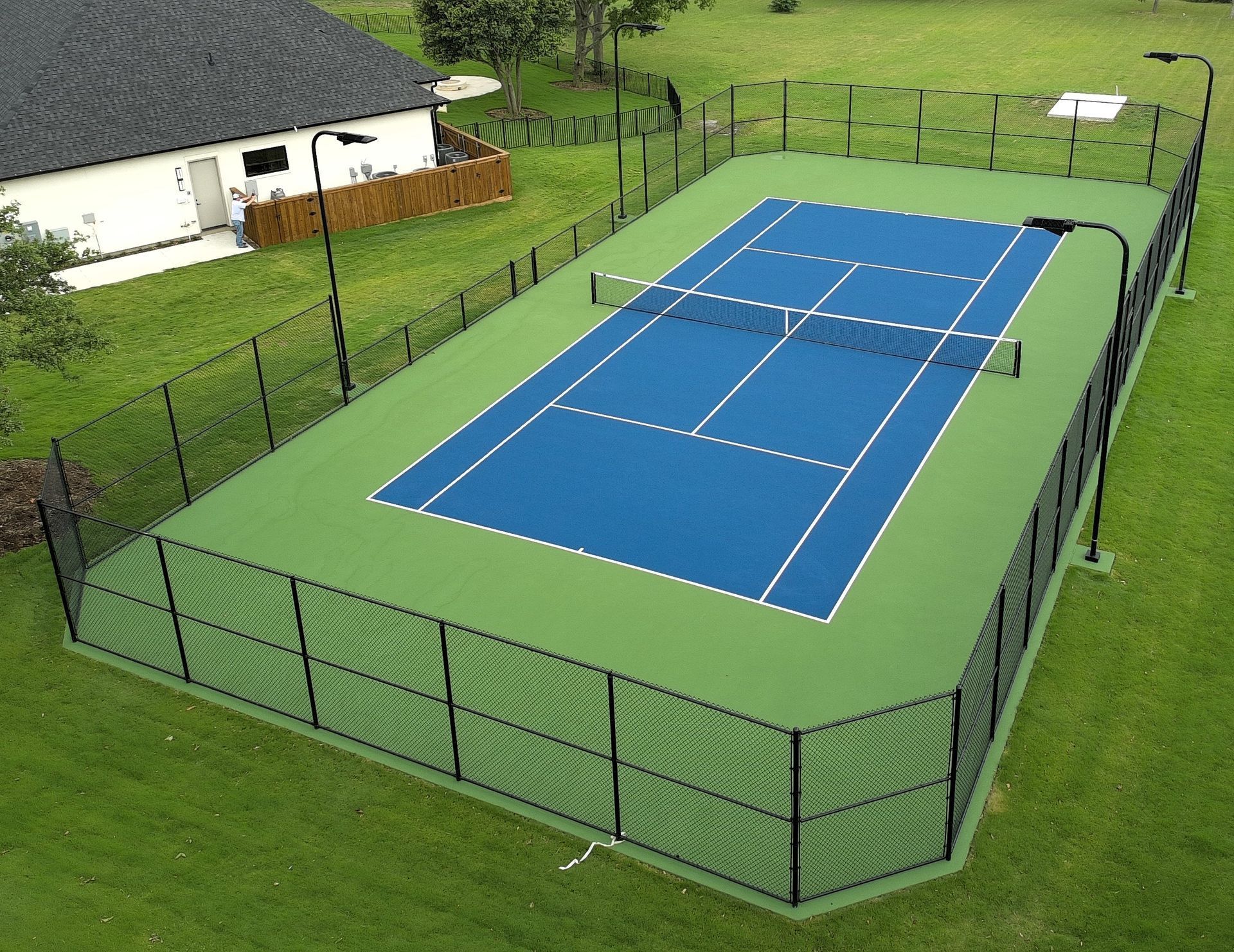 Fenced outdoor tennis court with blue playing surface and green surrounding area рядом