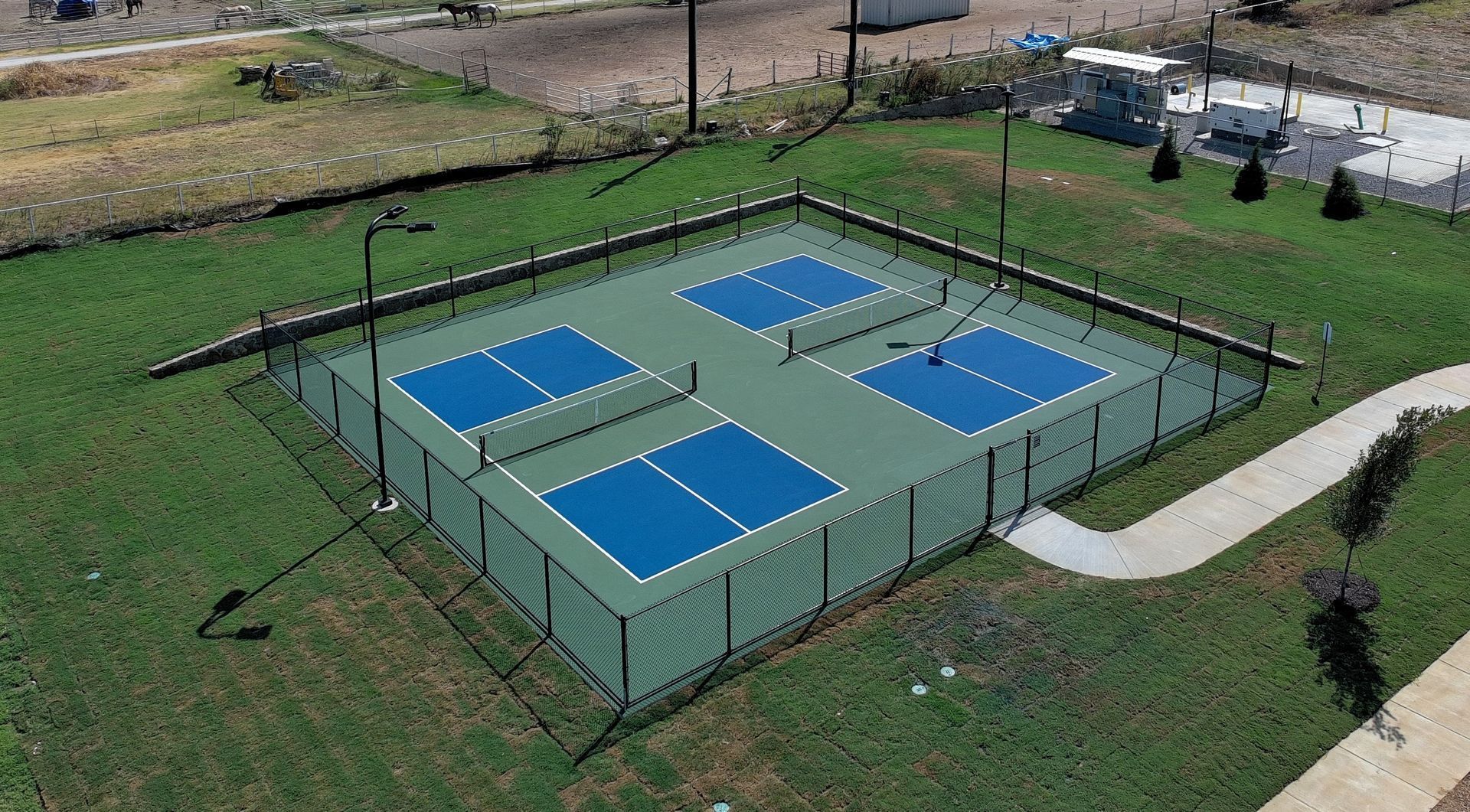Aerial view of a fenced outdoor pickleball court with four blue playing areas on a grassy field.