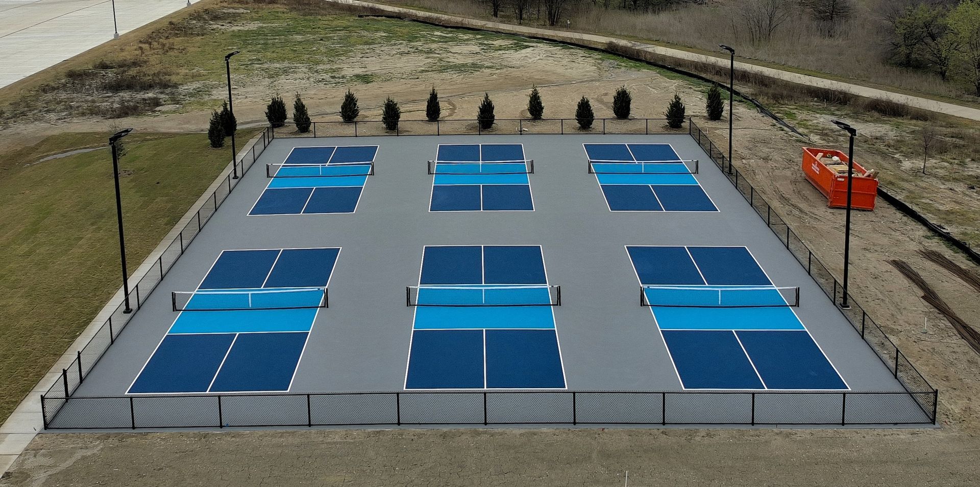 Overhead view of a public pickleball court with six playing areas, surrounded by a fence and trees.