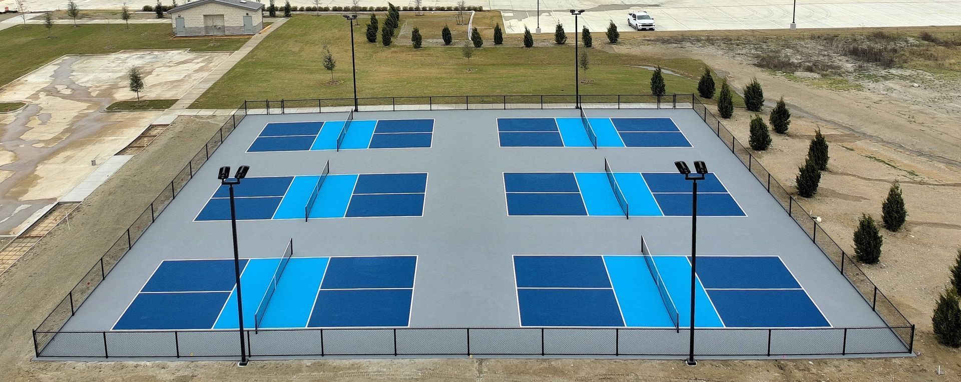 Aerial view of six pickleball courts, blue and gray, in a fenced area with trees and a building in the background.