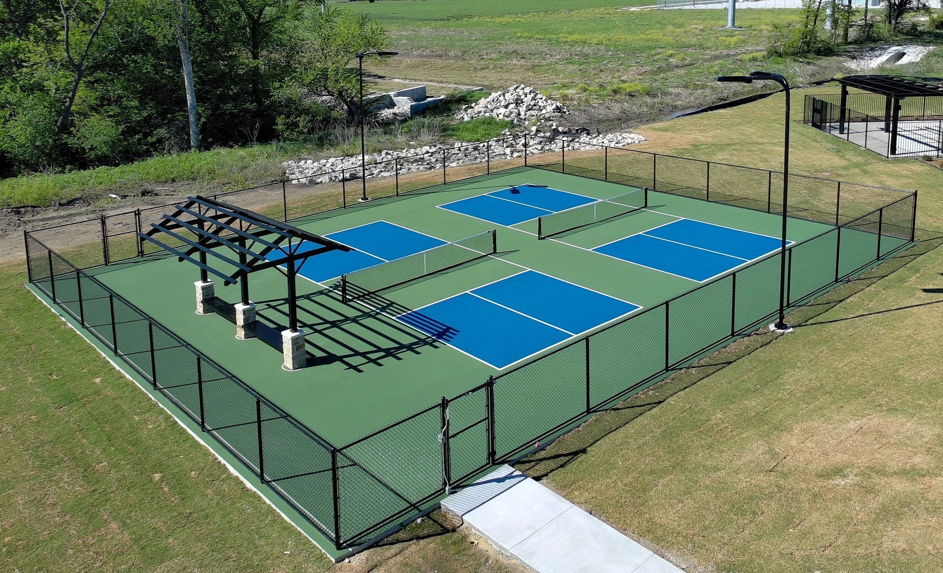 An aerial view of four pickleball courts with blue playing surfaces, a green perimeter, and a black fence, set on grass.