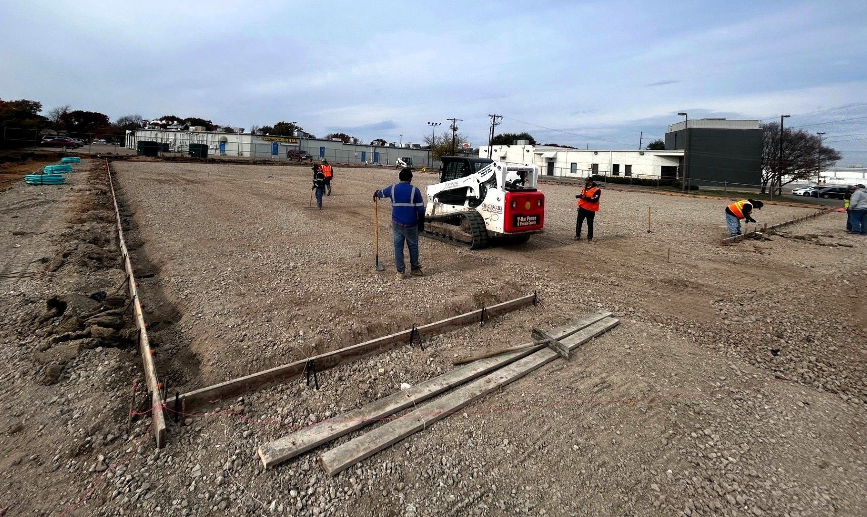 Construction workers grading gravel with a skid steer and hand tools on a construction site.