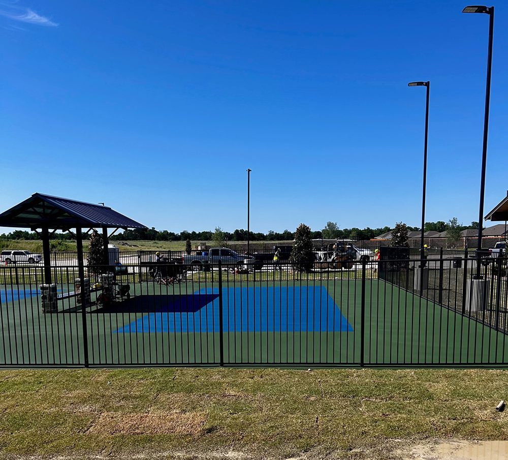 Fenced pickleball court with blue and green surface, shelter, poles, cars in background under a blue sky.