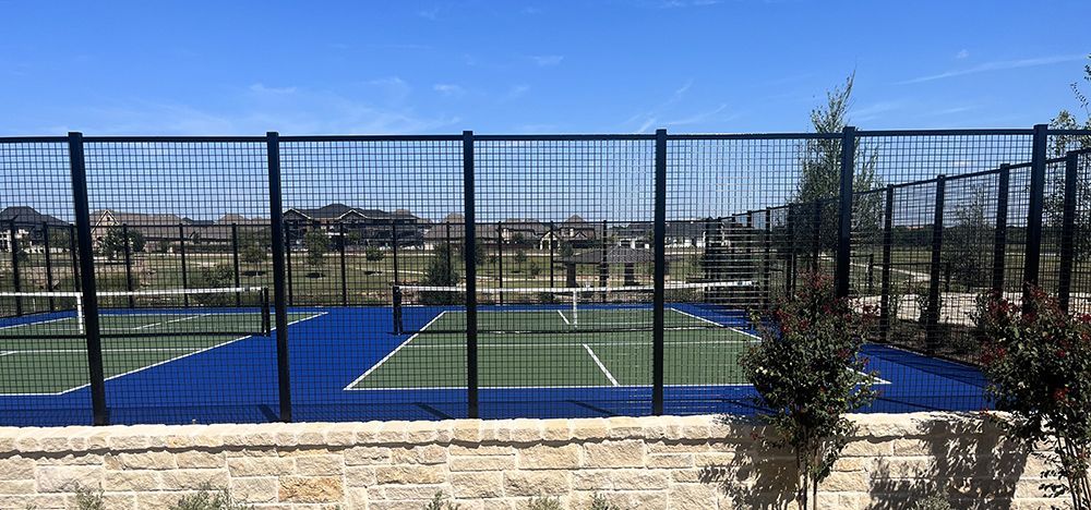 Tennis court behind a black wire fence, blue and green surface, sunny day.