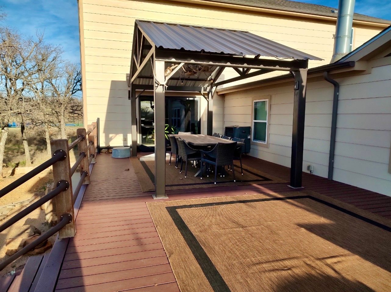 Deck with outdoor dining table under a pergola, adjacent to a house, brown wood and rug, blue sky.