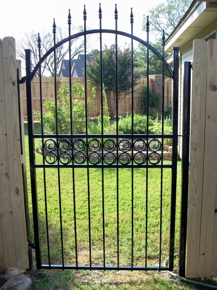 Black metal garden gate in a wooden fence; green grass and foliage visible.