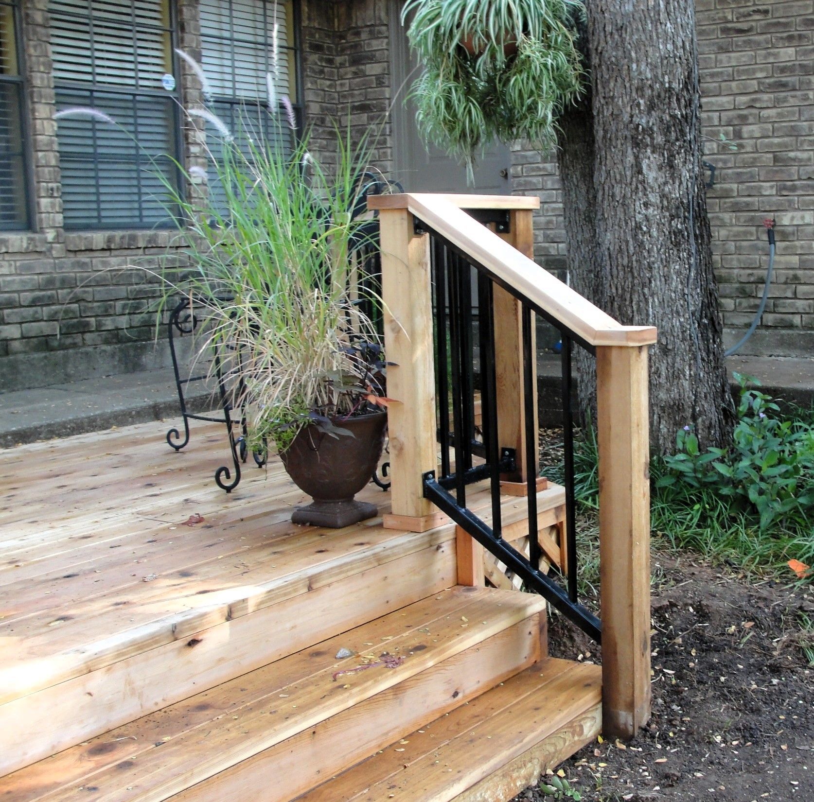 Wooden deck with steps, black metal handrail, potted plants. Brick house background.