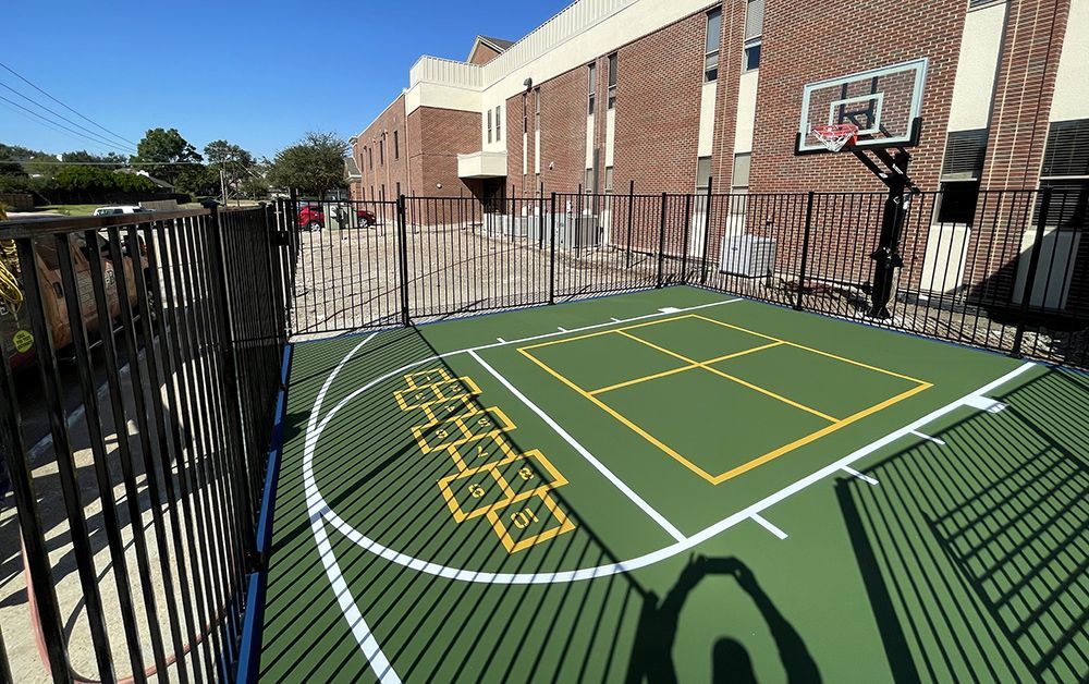 Small outdoor basketball court with green surface, black fencing, and brick building.