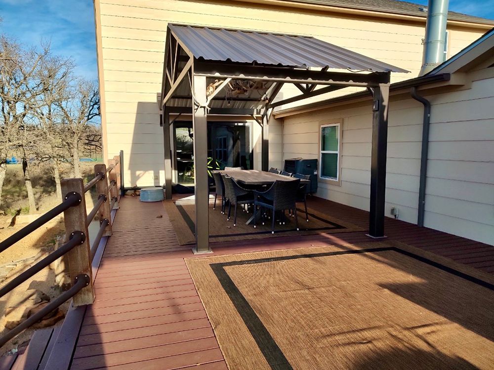 A covered patio with a table and chairs, on a wooden deck next to a house.