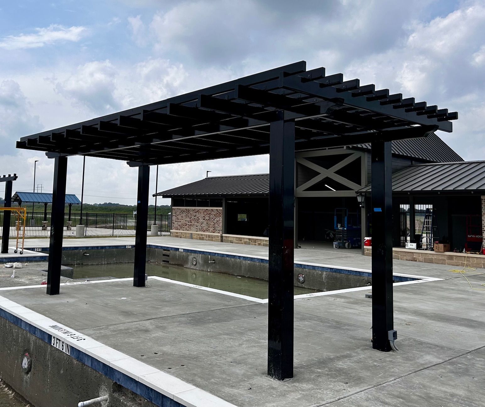 Black pergola over a concrete pool.  Building and overcast sky in the background.