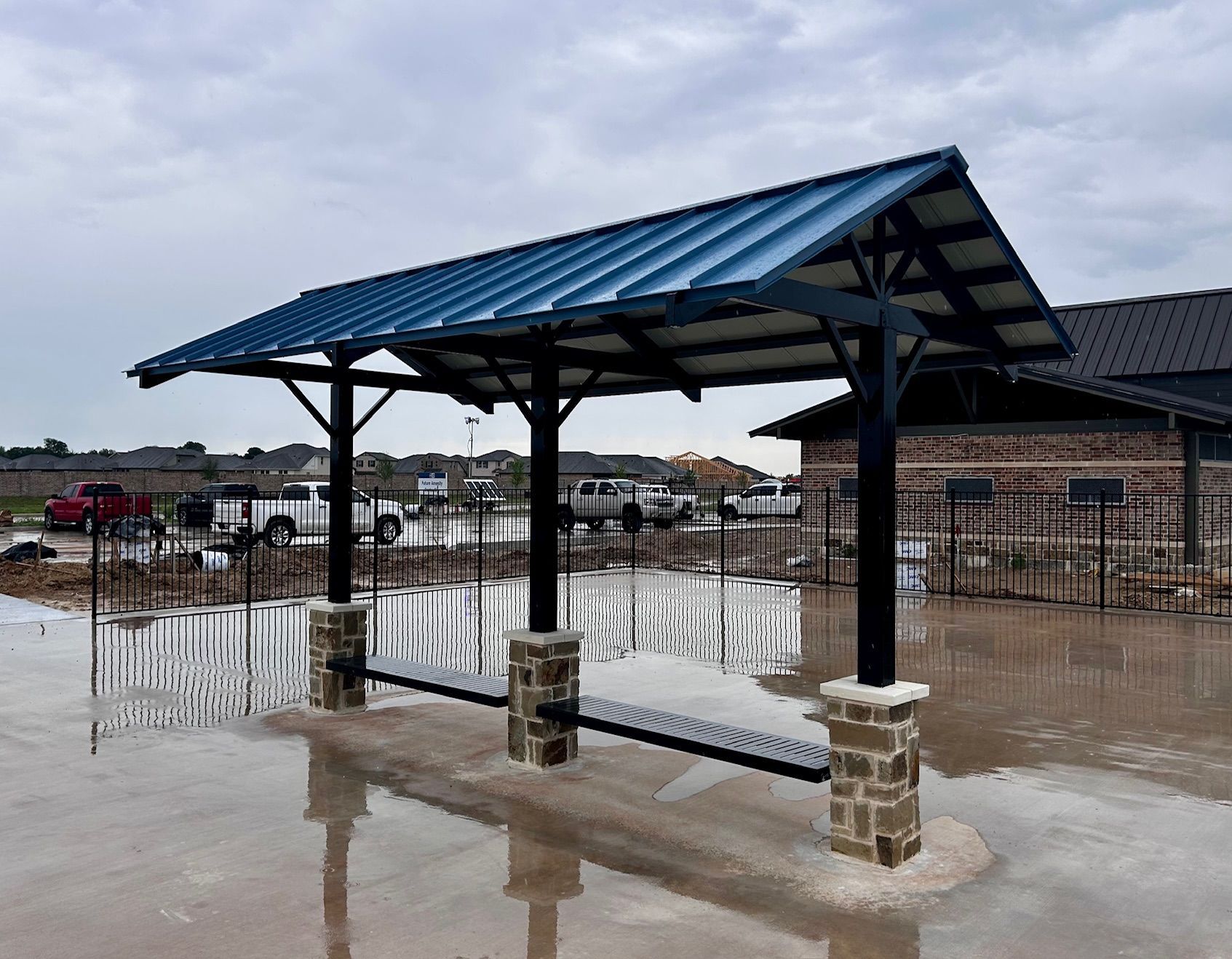 Covered shelter with dark blue roof, black supports, and stone columns; construction site in background.