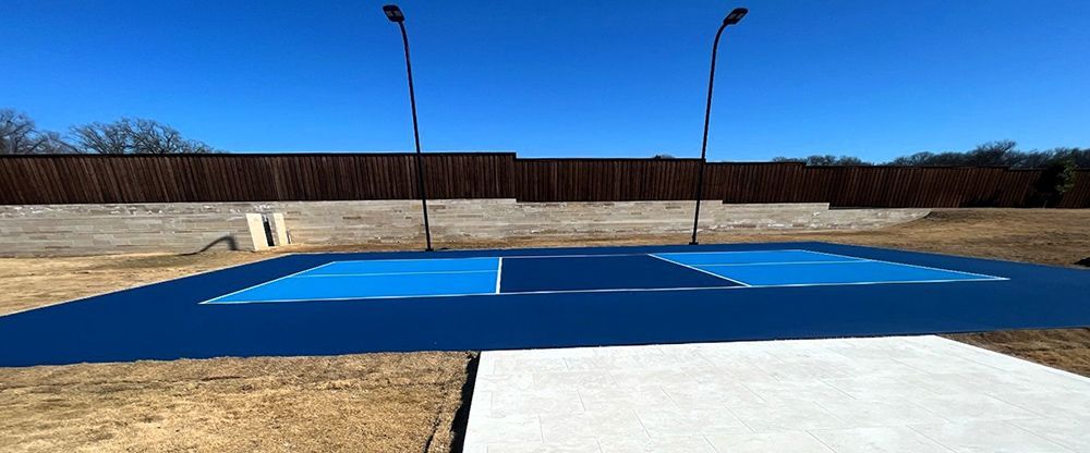 Blue pickleball court with two tall lights against a brown fence and a clear blue sky.