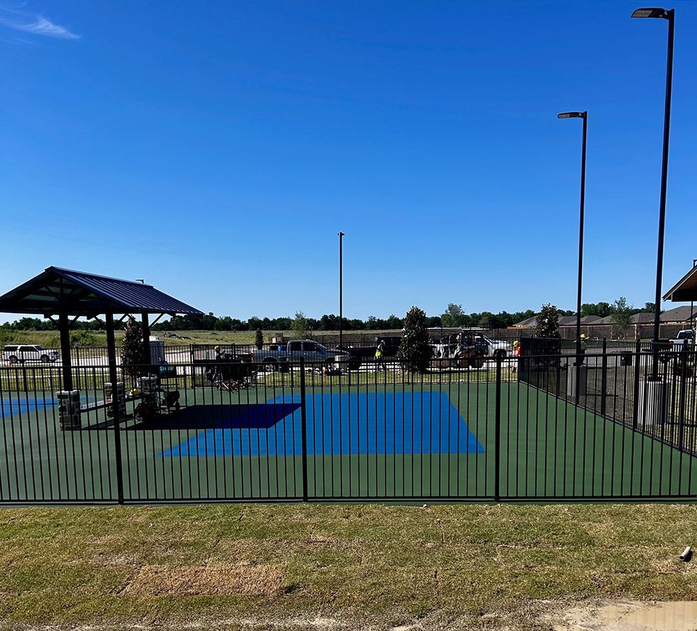Playground with blue and green ground, black fence, shaded area, and vehicles in the distance on a sunny day.