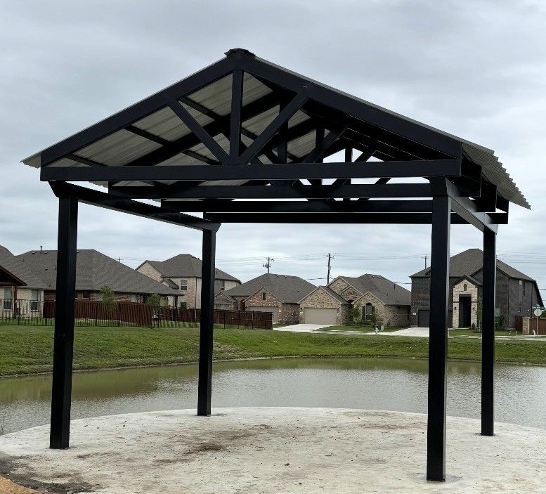 Black metal gazebo with a white roof over a concrete pad, overlooking a pond, residential houses in the background.