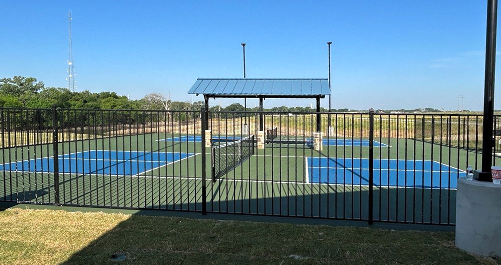 Pickleball courts behind a black fence. Blue playing surfaces, green grass, and a blue-roofed shelter. Sunny day.
