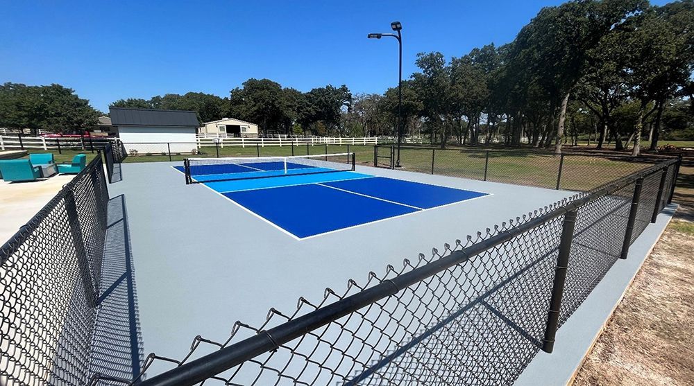 Pickleball court with blue and gray surface, black fence, and trees in the background.