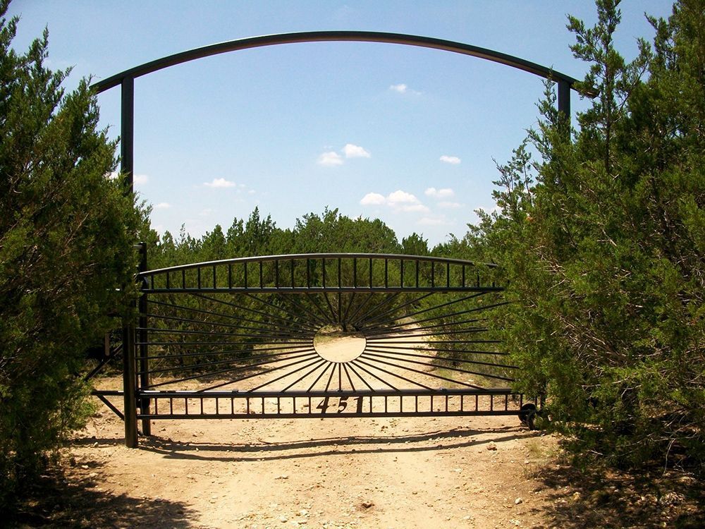 Black iron gate with sunburst design, arched top, set in a dirt driveway lined with green trees against a blue sky.